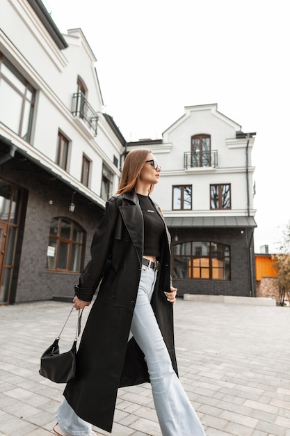 Modelo de mujer de belleza callejera de moda con traje negro de moda con jeans largos de abrigo de cuero y elegante bolso camina por la calle