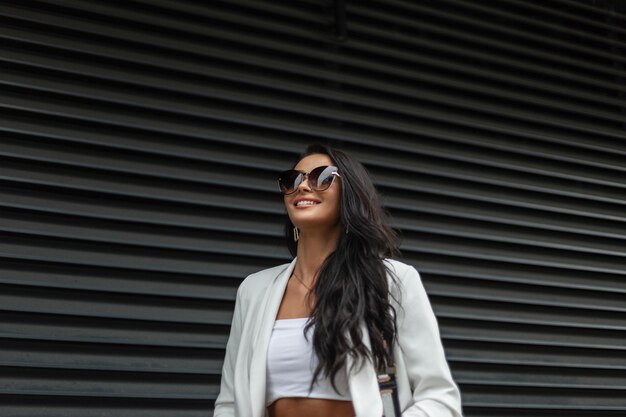 Modelo de mujer de belleza de negocios de moda con sonrisa con gafas de sol de moda en elegante atuendo casual blanco camina en la ciudad cerca de una pared negra de metal