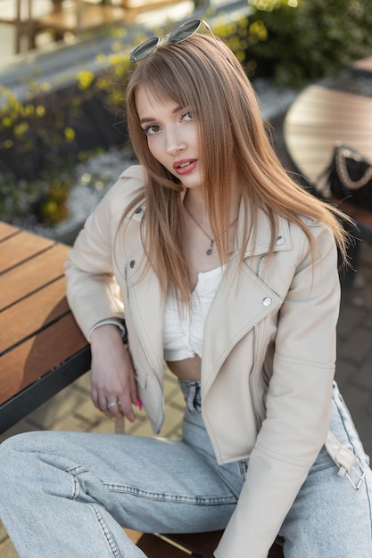 Modelo de mujer bonita fresca de moda con peinado en chaqueta de cuero con estilo de rock con camiseta y jeans vintage azules se sienta en un banco de madera y mira a la cámara Traje de estilo femenino de la calle