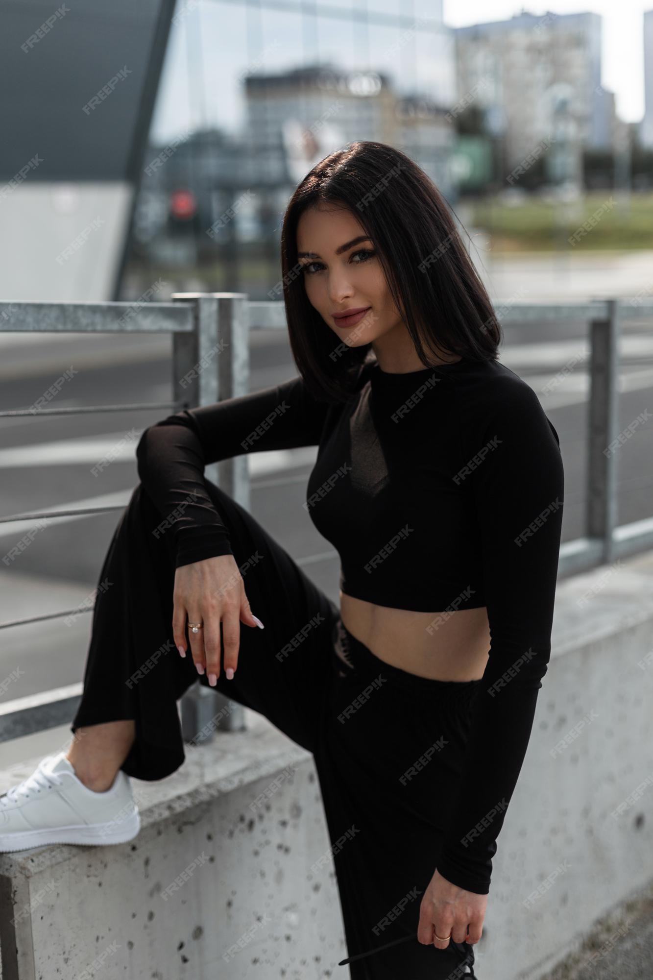 Modelo mujer bonita joven y elegante con ropa deportiva negra moda con pantalones de manga larga y zapatillas blancas posa en la ciudad | Foto Premium