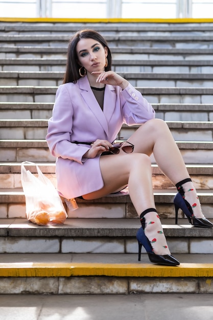 Modelo de mujer bonita de moda con naranjas de frutas se sienta en los escalones con una chaqueta morada, zapatos negros.