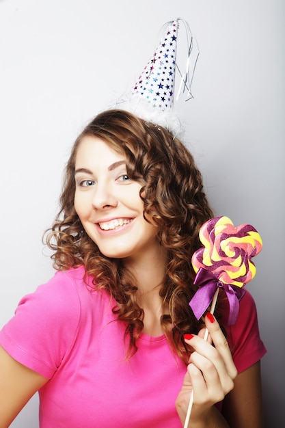 Modelo de mujer hermosa fiesta con piruleta, con sombrero de papel sobre fondo blanco.