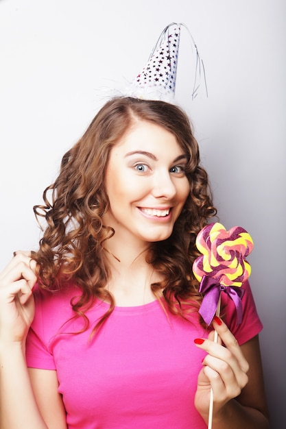 Modelo de mujer hermosa fiesta con piruleta, con sombrero de papel sobre fondo blanco.