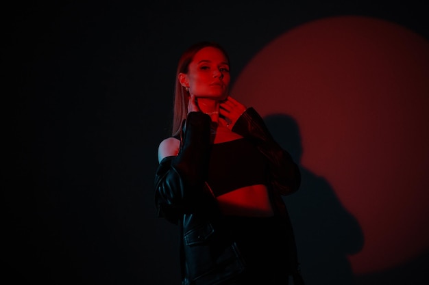 Modelo de mujer hermosa de moda en ropa negra elegante con chaqueta de cuero sobre fondo oscuro en estudio con luz creativa roja