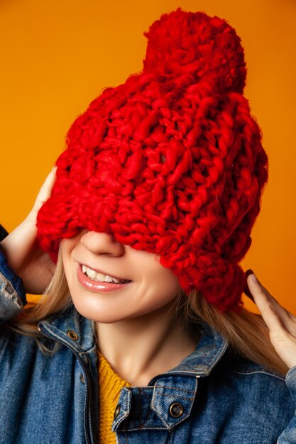 Modelo de mujer joven en chaqueta de mezclilla casual y gorro de punto rojo que cubre la mitad de la cara de pie contra el fondo naranja