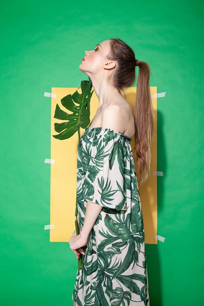 Modelo de mujer joven en elegante vestido con adornos florales sosteniendo la hoja de monstera mientras está de pie contra la hoja de papel amarillo sobre fondo verde en verano