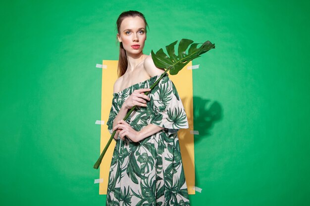 Modelo de mujer joven en elegante vestido con adornos florales sosteniendo la hoja de monstera mientras está de pie contra la hoja de papel amarillo sobre fondo verde en verano