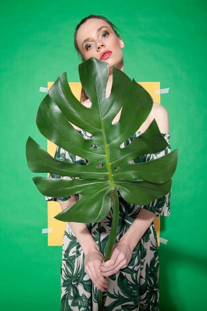 Modelo de mujer joven en elegante vestido con adornos florales sosteniendo la hoja de monstera mientras está de pie contra la hoja de papel amarillo sobre fondo verde en verano