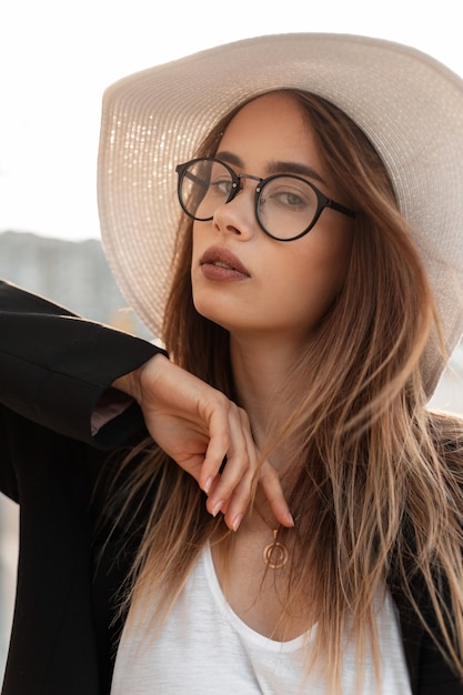 Modelo de mujer joven encantadora con estilo en gafas de moda en sombrero de paja de moda en poses de chaqueta elegante negro de moda en la calle. Retrato fresco hermosa chica en ropa hermosa de moda al aire libre en la ciudad.