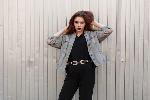 Modelo de mujer joven hermosa de moda en una elegante chaqueta y polo negro posa cerca de una pared de metal en la calle