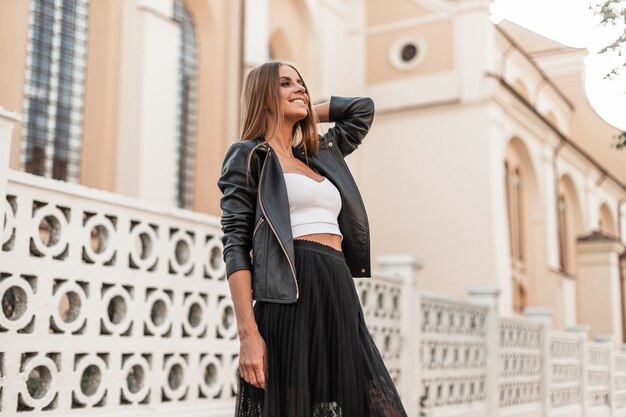 Foto modelo de mujer joven muy feliz en ropa elegante y elegante está de pie y linda sonriendo cerca de una valla vintage en la ciudad en un cálido día de otoño. hermosa chica alegre descansa en la calle. estilo.