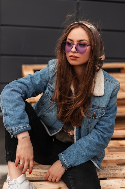 Modelo de mujer muy de moda estadounidense en elegantes gafas púrpuras en chaqueta de mezclilla azul vintage descansa sobre paletas de madera cerca de un edificio moderno en la ciudad. Hermosa chica hipster. Moda casual.