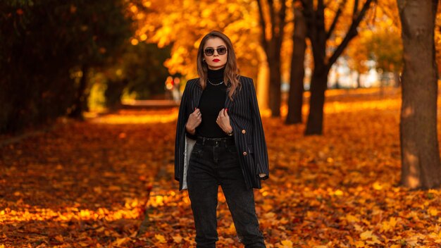 Modelo de mujer de negocios fresca con elegantes gafas de sol en ropa de traje negro de moda con blazer de moda camina en el parque de otoño con follaje naranja