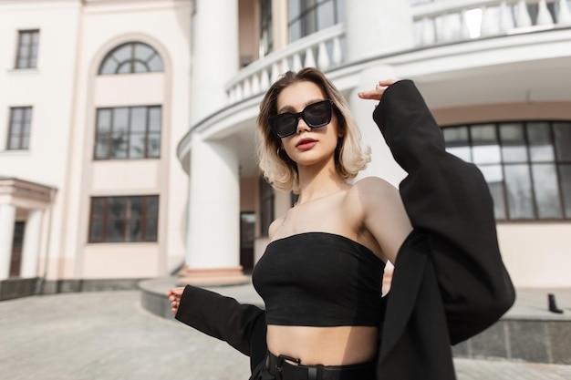 Modelo de mujer de negocios joven de moda con elegantes gafas de sol modernas en ropa elegante de moda negra con top y blazer camina en la calle cerca de un edificio antiguo Estilo urbano y moda