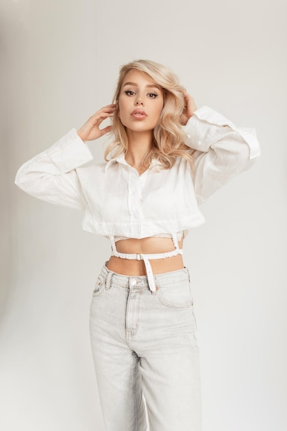 Modelo de mujer sexy glamurosa con cabello rubio en un moderno atuendo casual con una camisa y jeans sobre un fondo blanco en el estudio