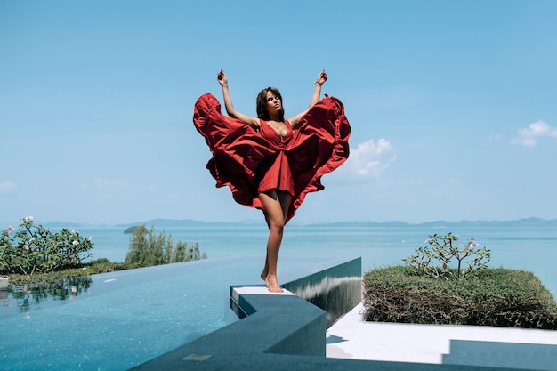 Modelo de mujer en vestido rojo de moda de pie al borde de la piscina infinita con vistas al mar.