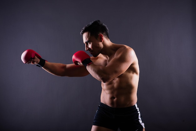 Modelo musculoso deportivo joven en guantes de boxeo sobre fondo gris macho flexionando sus músculos