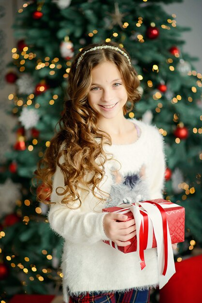 Modelo de niña hermosa cerca del árbol de Navidad tiene un regalo de año nuevo en una caja roja. Retrato de primer plano