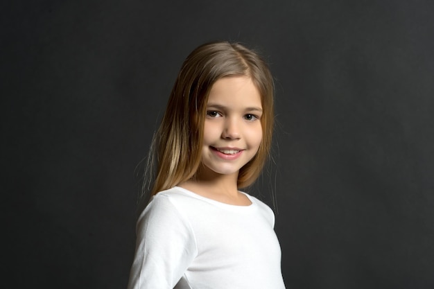 Modelo de niño sonriendo con cabello largo sano. Chica con sonrisa en cara adorable sobre fondo oscuro. Belleza, mirada, peinado. Niño feliz, concepto de infancia. Juventud, cuidado de la piel, salud.