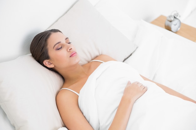 Modelo de pelo marrón joven natural en pijama blanco durmiendo en su cama