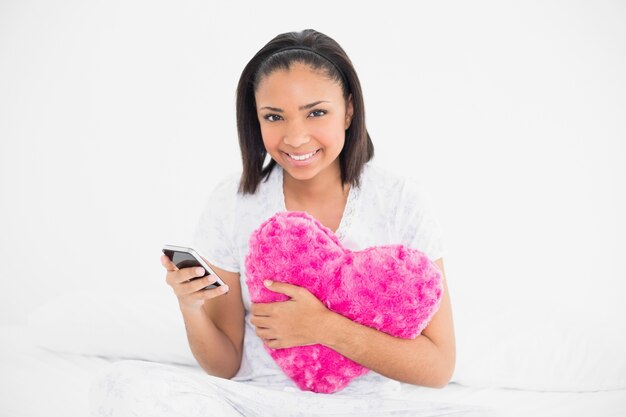 Modelo de pelo oscuro joven encantado sosteniendo una almohada y un teléfono móvil