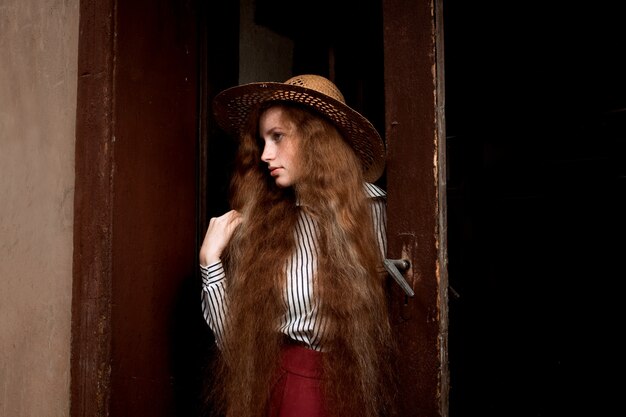 Modelo de pelo rojo joven atractivo con el pelo largo y ondulado en el sombrero de paja que presenta cerca de la puerta vieja