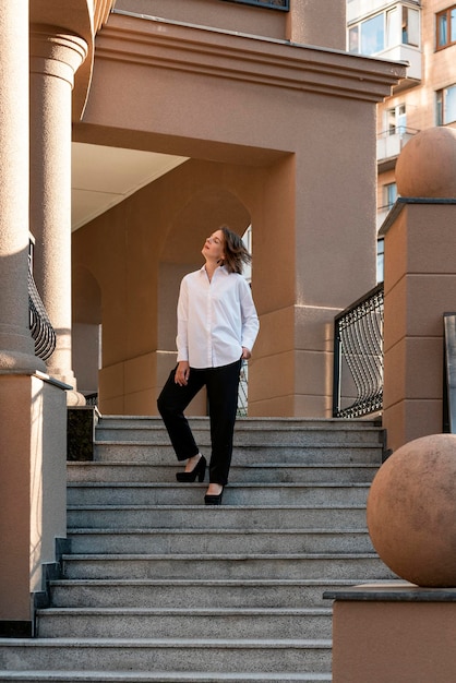 Modelo pensativamente posa nos degraus do edifício arquitetônico Garota está vestida com camisa e calças de estilo clássico