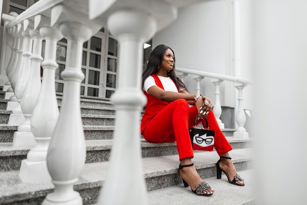 Modelo positivo da mulher jovem africana em roupa elegante vermelha com bolsa em sapatos da moda sorri e descanse na escada de pedra vintage na cidade perto do prédio. feliz garota negra em terno da moda ao ar livre