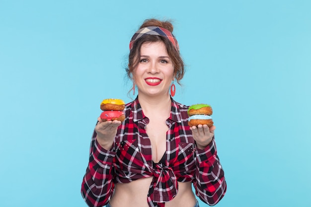 Modelo positivo de jovem pin-up segurando nas mãos quatro donuts multicoloridos posando sobre um fundo azul. Conceito de cozinha para sobremesas e doces. Espaço de publicidade