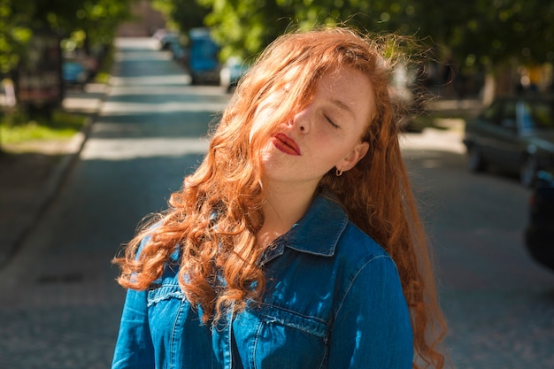 Modelo ruiva bonita em uma camisa jeans sob os raios de sol com os olhos fechados