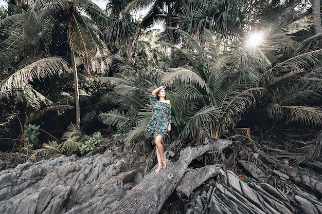 Modelo ruivo atraente em um vestido com estampa floral se passando perto de grandes palmeiras e de pé em uma velha árvore. Brilho do sol. Phuket. Tailândia.