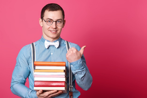 Modelo sincero positivo mira directamente a la cámara con una enorme pila de libros con una mano