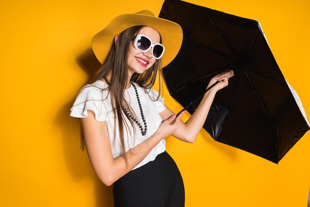 Modelo sorridente de garota de cabelos compridos confiante com chapéu e óculos de sol segura guarda-chuva e posando em fundo amarelo