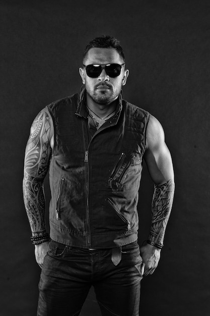Modelo de tatuaje con barba en la cara sin afeitar Hombre barbudo con tatuaje en brazos fuertes Hombre tatuado con bíceps y tríceps Macho de moda con gafas de sol de moda Estilo de moda y filtro vintage de tendencia