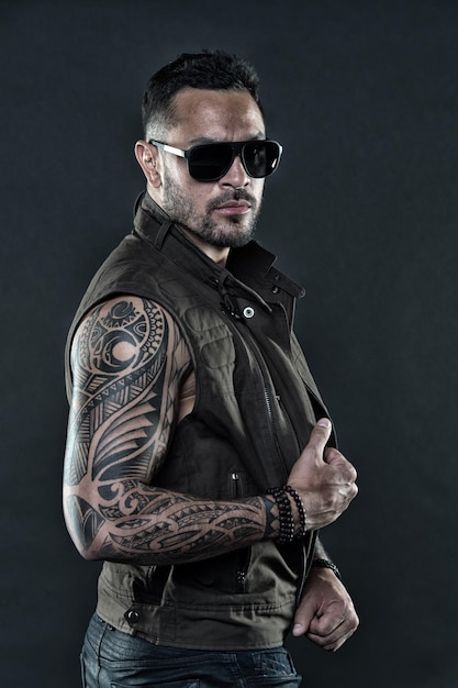 Modelo de tatuaje con barba en la cara sin afeitar Hombre barbudo con tatuaje en brazos fuertes Hombre tatuado con bíceps y tríceps Macho de moda con gafas de sol de moda Estilo de moda y filtro vintage de tendencia