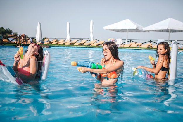 Modelos alegres y divertidos jugando en la piscina. Sostienen pistolas de agua en las manos y lo usan. Dos mujeres están en contra de la tercera. Ellos sonríen y ríen