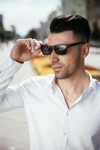 Modemann in einem weißen Hemd steht an einem sonnigen Tag auf der Straße und posiert und sieht zu, wie er eine Brille hält