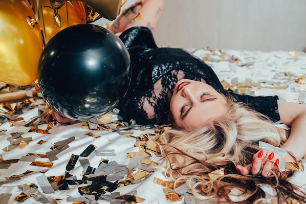 Modemodellbild. Nach der Party Entspannung. Blonde Frau, die mit geschlossenen Augen auf dem Bett liegt. Luftballons und Konfetti herum.