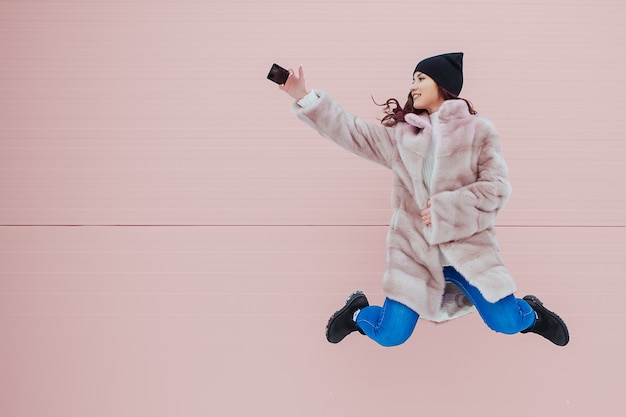 Modeporträt der hübschen lächelnden und springenden Frau im Nerzmantel mit Smartphone gegen die bunte rosa Wand. Copyspace.