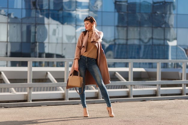 Modeporträt der jungen vorbildlichen Frau in schönem braunem beige Mantel, Denim-Jeans und Sonnenbrille auf städtischem Hintergrund.