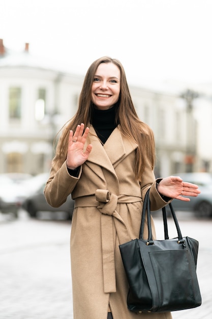 Modeporträt der stilvollen jungen Frau, die Spaß, emotionales Gesicht, lachend hat. Urban City Street Style mit dunklen Freizeithosen, weißem Pullover, cremigem Mantel und Sonnenbrille. Modekonzept.