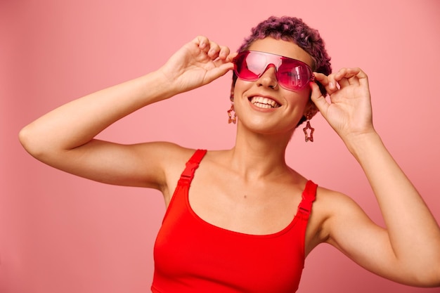 Modeporträt einer Frau mit kurzem Haarschnitt, farbiger Sonnenbrille und einem Lächeln mit Zähnen in einem roten Top auf rosa Hintergrund