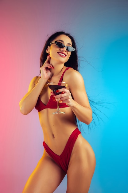 Modeporträt einer jungen, fitten und sportlichen Frau mit Cocktail in stilvoller roter Luxusbadebekleidung an einer Wand mit Farbverlauf, perfekter Körper für den Sommer bereit summer