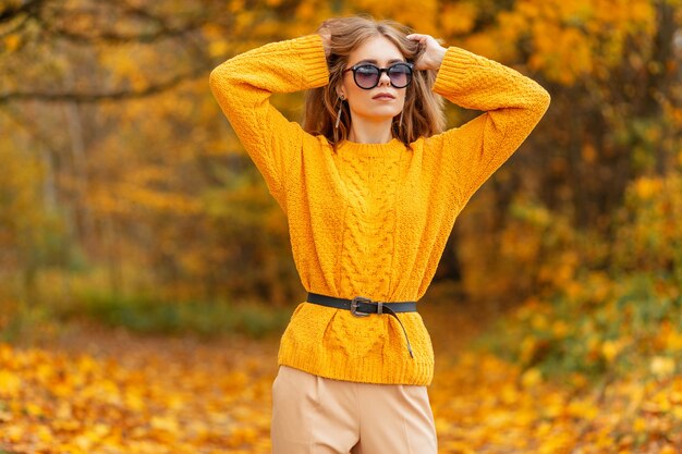 Modeporträt einer schönen Frau mit stilvoller Sonnenbrille in einem gelben Strickpullover im Vintage-Stil geht an einem Herbsttag in der Natur mit buntem gelbem Herbstlaub