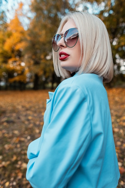 Modeporträt einer schönen jungen blonden frau mit stylischer sonnenbrille in einem modischen blauen mantel auf natur auf einem hintergrund von gelbem herbstlaub