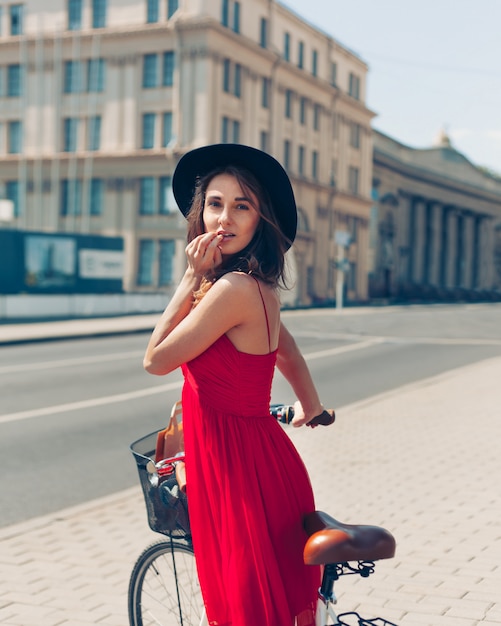 Modeporträt im Freien des attraktiven jungen Brunette in einem Hut auf einem Fahrrad.