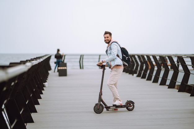 Modern Junger Mann mit Elektroroller auf einem Pier in der Nähe des Meeres Ökologisches Transportkonzept