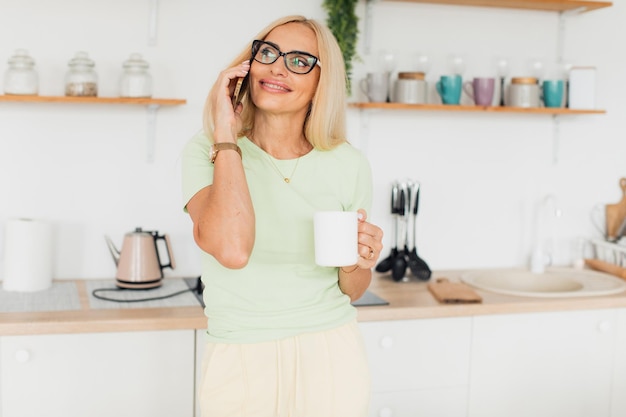 Moderna y atractiva mujer de mediana edad hablando por teléfono y bebiendo café en la cocina de casa