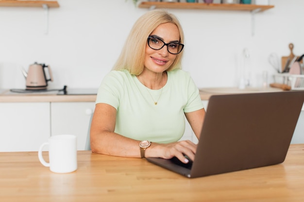 Moderna y atractiva mujer de mediana edad tomando café y trabajando en una laptop en la cocina de casa