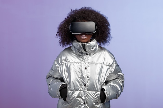 La moderna chica de cabello castaño rizado vestida con una chaqueta plateada usa las poses de gafas de realidad virtual en el estudio sobre fondo de neón.